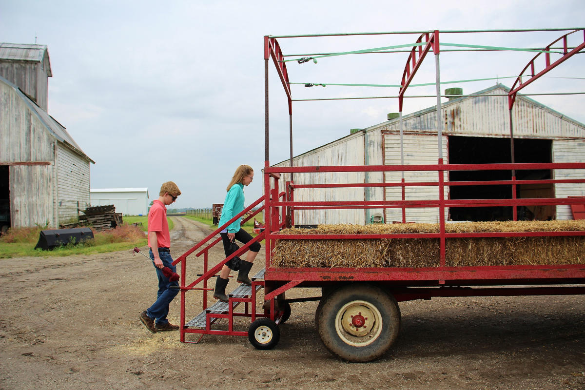 A teen boy and girl walk up the steps of a trailer with bales of hay.
