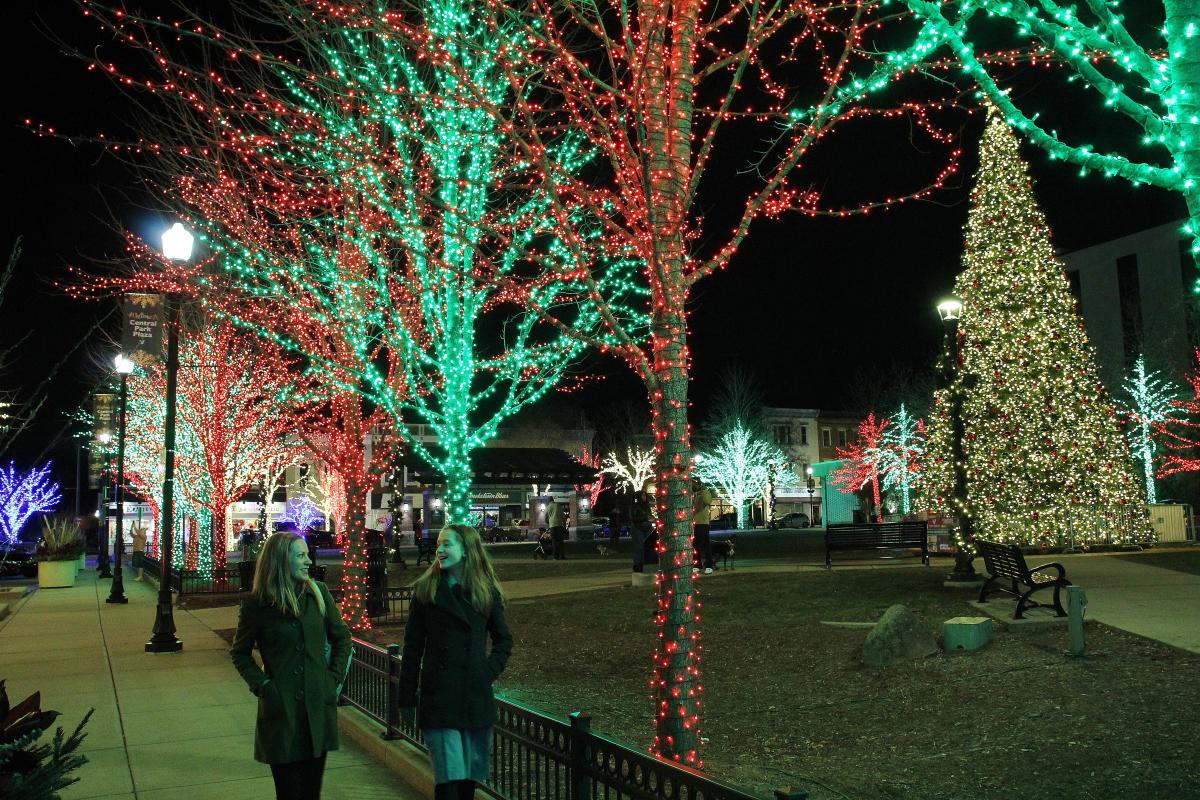A festive outdoor scene at night with trees wrapped in red and green holiday lights. A large Christmas tree covered in twinkling white lights stands to the right, while additional trees with blue, white, and red lights line the background. Two women in winter coats walk along a sidewalk, smiling at each other. Behind them, a lit pavilion and nearby buildings contribute to the lively, illuminated atmosphere of the holiday season.