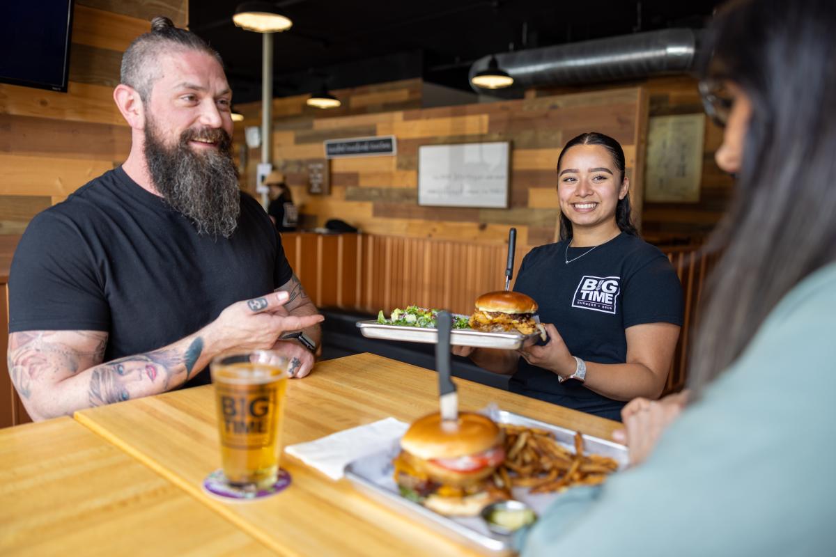A waitress holds a plate with a burger on it in front of a table with two customers.