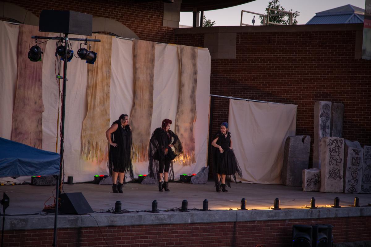 Three people in dark costumes stand on an outdoor stage. Tan and white striped cloth hangs in the back. Tall stones with symbols on them stand to the right.