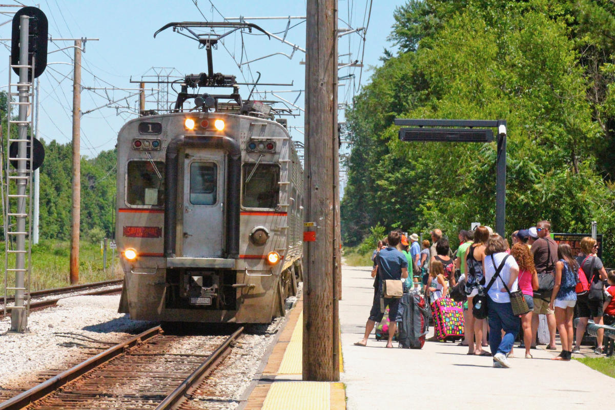 A commuter train stopped at a station with riders standing on the platform to the right.