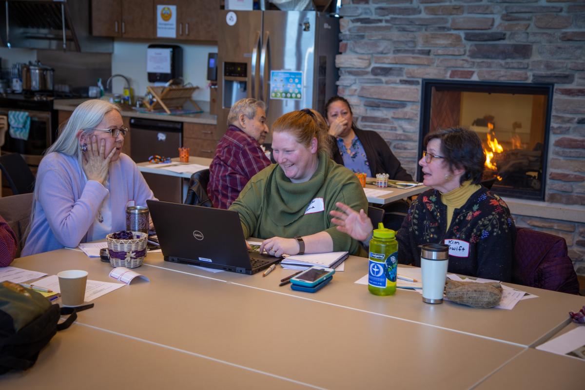 Three women site in front of a laptop on a table, smiling. Two people sit at an additional table in the background, near a fireplace.