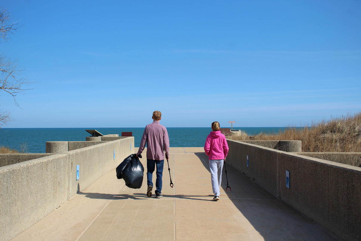 Two people walk toward the lake in the distance. One is holding a black trash bag and a trash grabber.