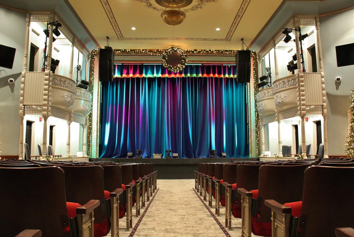 An elegant theater with a view from the audience seating area looking toward the stage. Rows of dark wooden chairs with red cushions line the aisle, leading up to a stage framed by ornate white balconies. The stage curtains are deep blue with soft pink and purple lighting. A wreath decorated with twinkling lights hangs above the stage, and garlands adorn the proscenium, adding a festive touch. The ceiling features intricate molding and a golden chandelier.