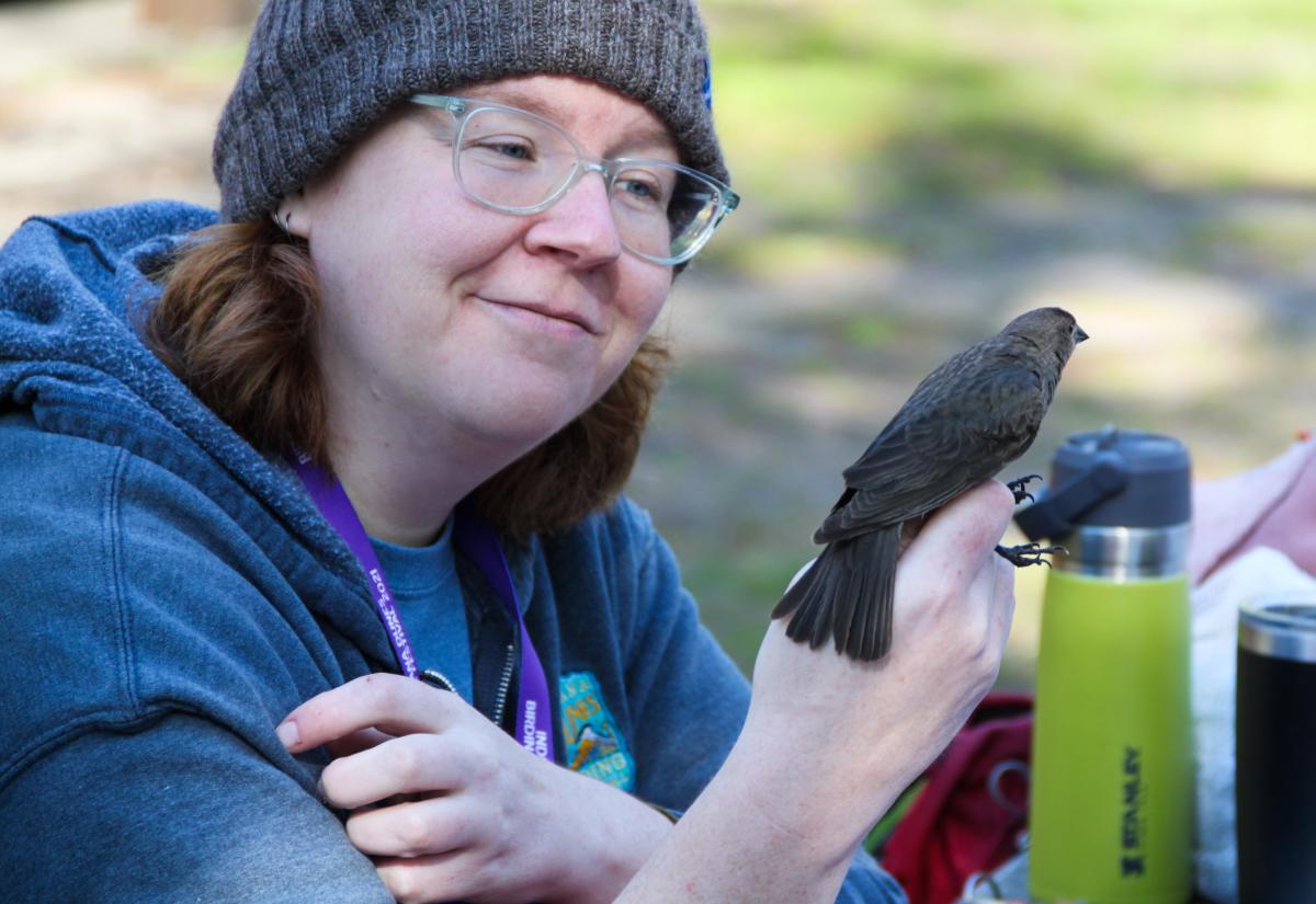 A person with glasses and a beanie enjoys a quiet moment with a small brown bird perched on their hand. The relaxed setting, with thermoses and bags in the background, suggests a nature excursion or birdwatching experience.