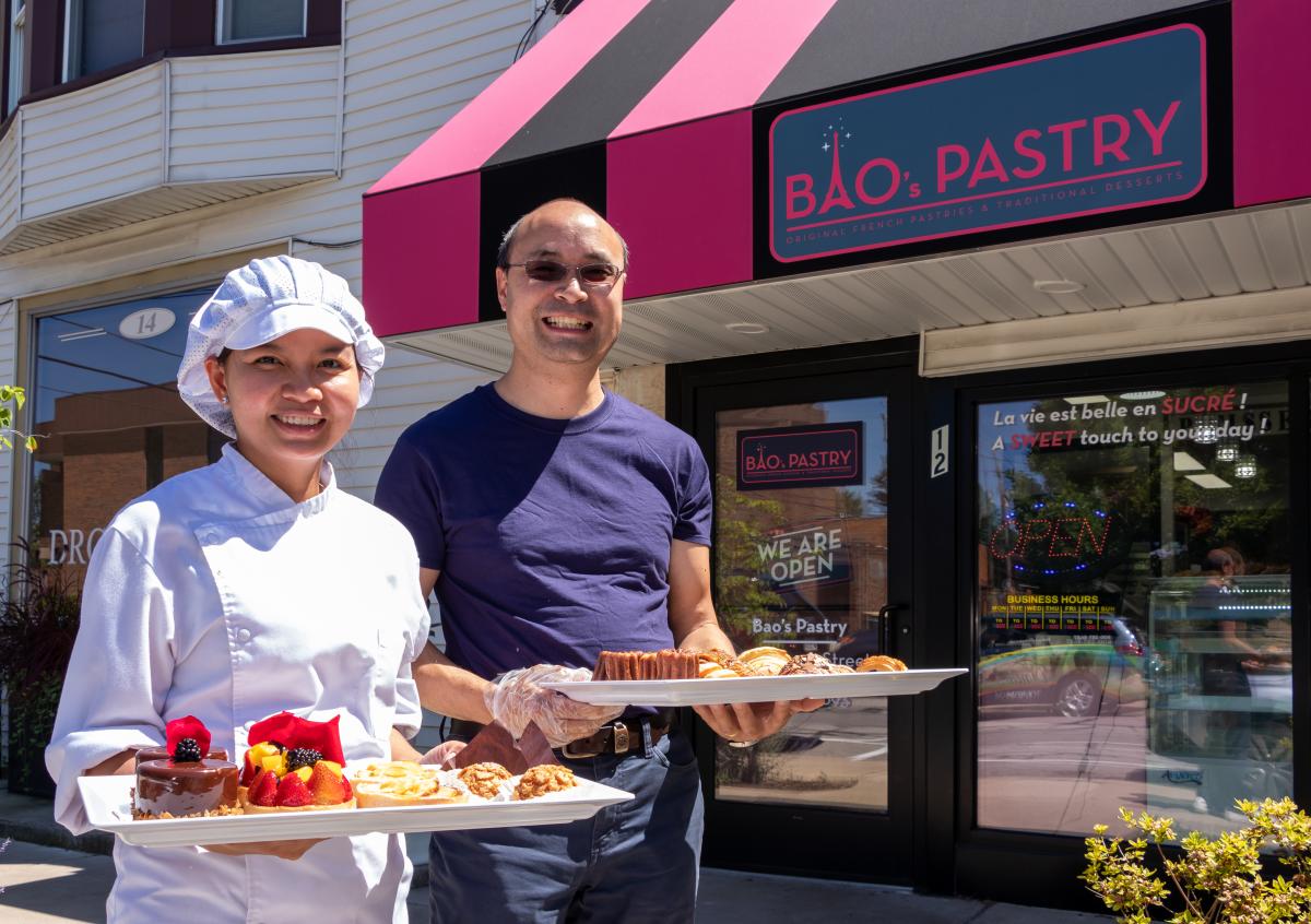 A chef and her husband stand in front of their pastry shop, holding trays of French pastries