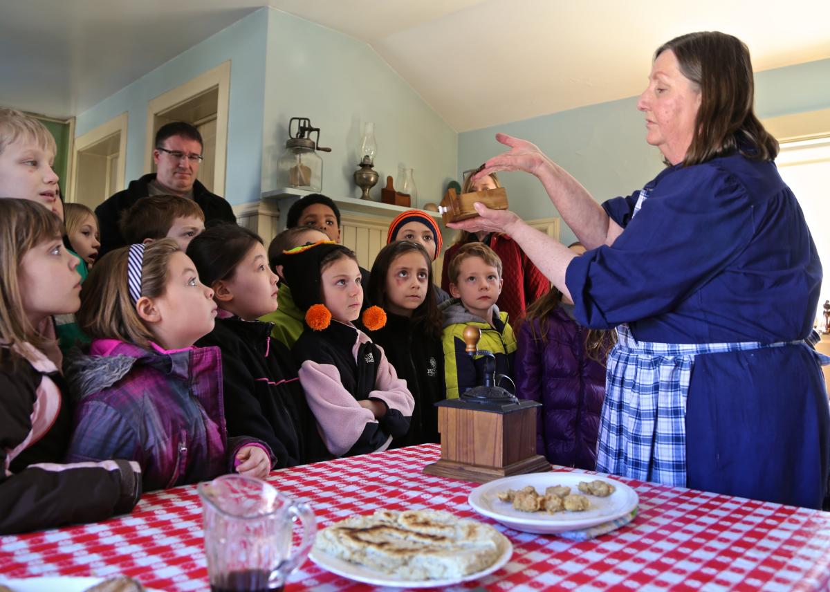 A class of school children look at a woman dressed in period clothes as she gives a talk about maple syrup products.