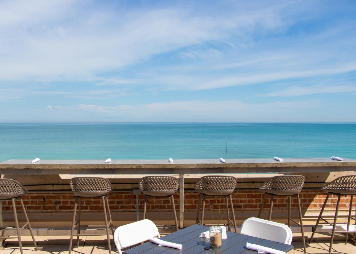 An outdoor lakeside dining deck at the Pavilion at Indiana Dunes, featuring bar stools and tables overlooking a sweeping view of Lake Michigan under a bright blue sky.