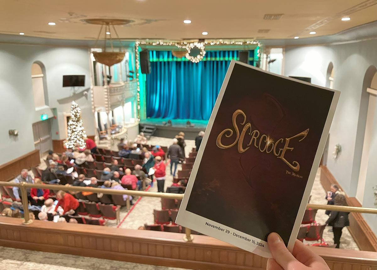 A person holds up a program for Scrooge: The Musical in a theater balcony, with the stage and audience in the background. The program cover has gold lettering on a dark background, and the dates "November 29 - December 15, 2024" are printed at the bottom. The theater is decorated with festive garlands and a lit Christmas tree. The stage has blue curtains, and the audience is filling the seats, some engaged in conversation.