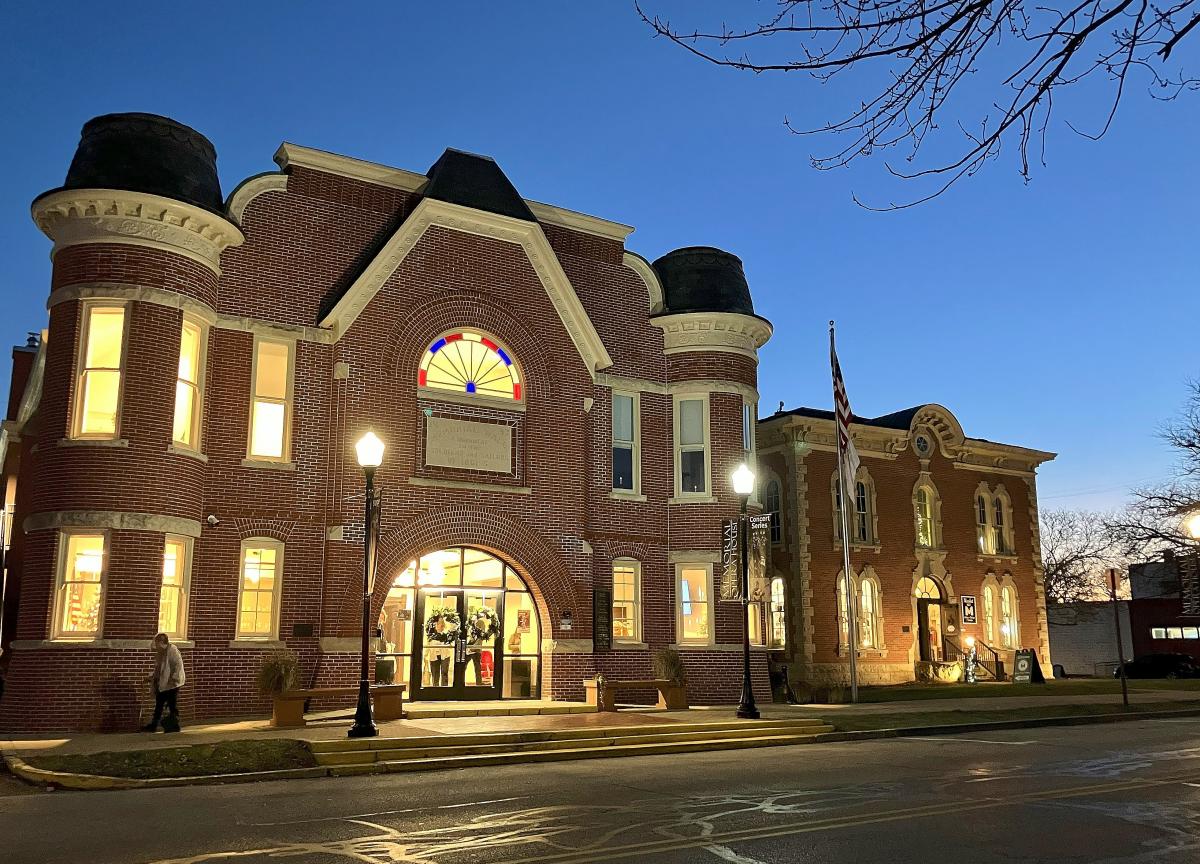 A historic red brick building with arched windows and decorative turrets is illuminated against the deep blue twilight sky. The entrance, framed by a large arched doorway, has glass doors adorned with festive wreaths. A second, similarly styled brick building with an American flag stands nearby. Street lamps cast a warm glow on the sidewalk, where a person in a coat walks along the edge of the frame.