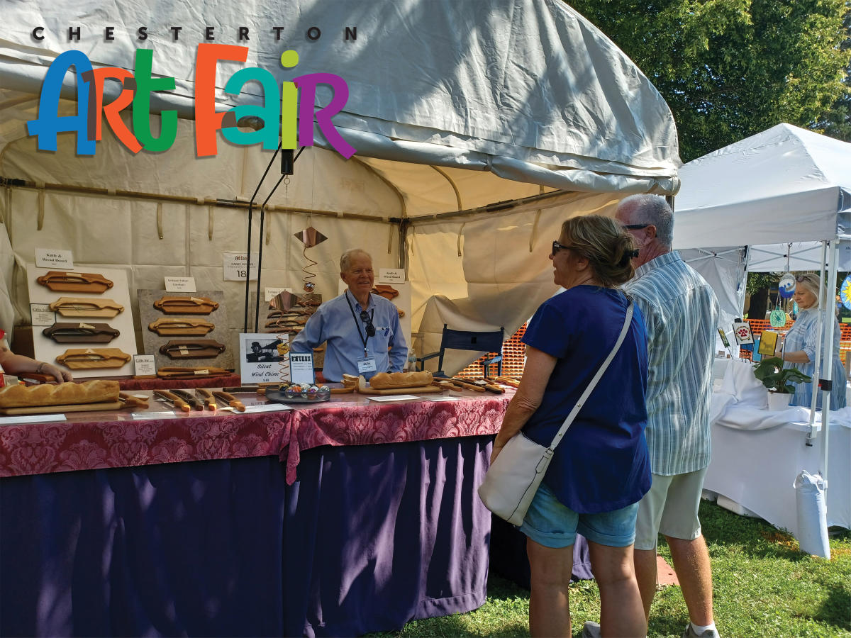 A vendor stands behind a table displaying bread boards and knives, talking to two customers at an outdoor art fair.