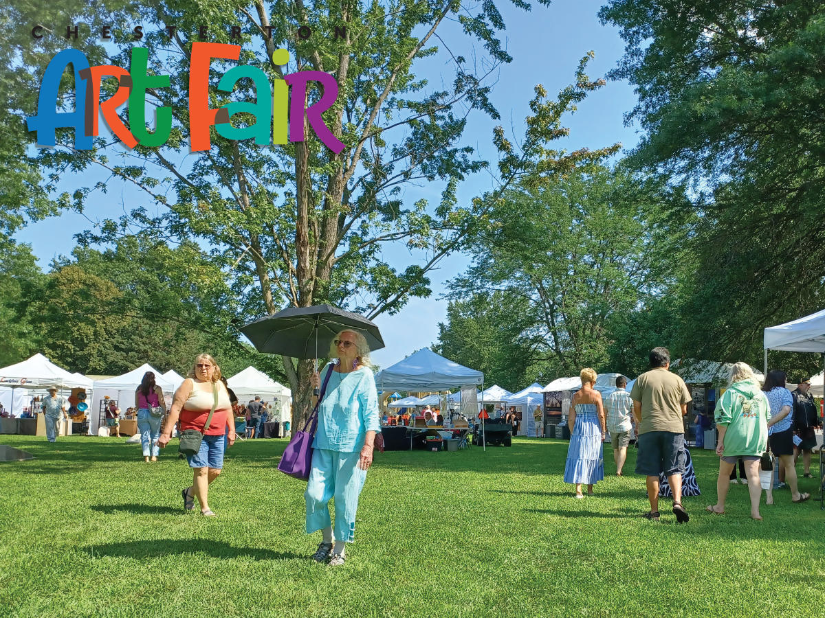 People walk through a sunny outdoor art fair with white tents and green trees.