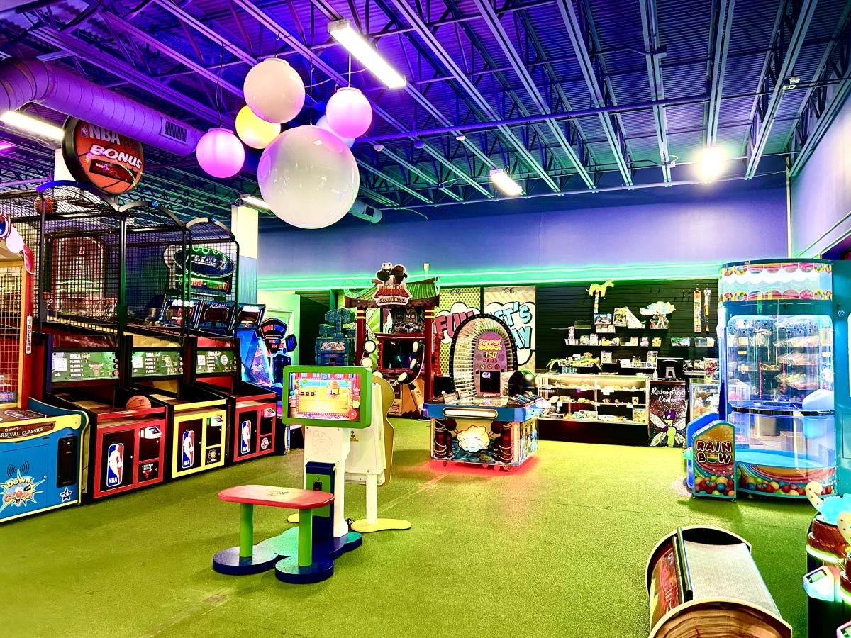 An arcade with a green carpet floor, several colorful games, and illuminated ceiling spheres under a blue metal roof.