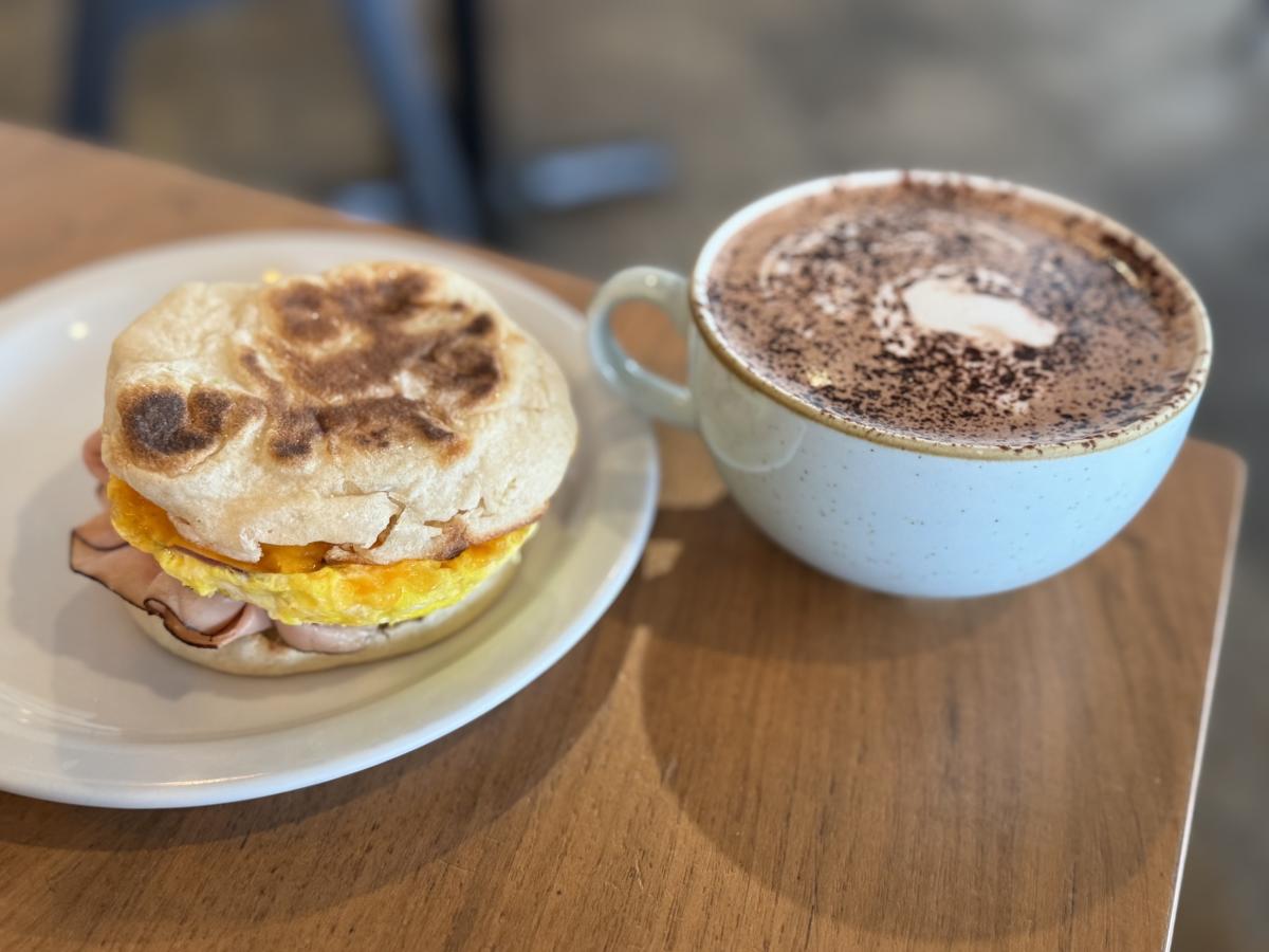Breakfast sandwich with egg, ham, and cheese on an English muffin, next to a cup of hot chocolate.