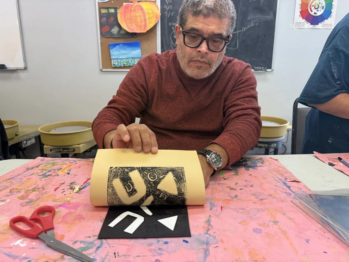 A man in a red sweater holds up a printed paper showing black and white abstract geometric art in a studio.