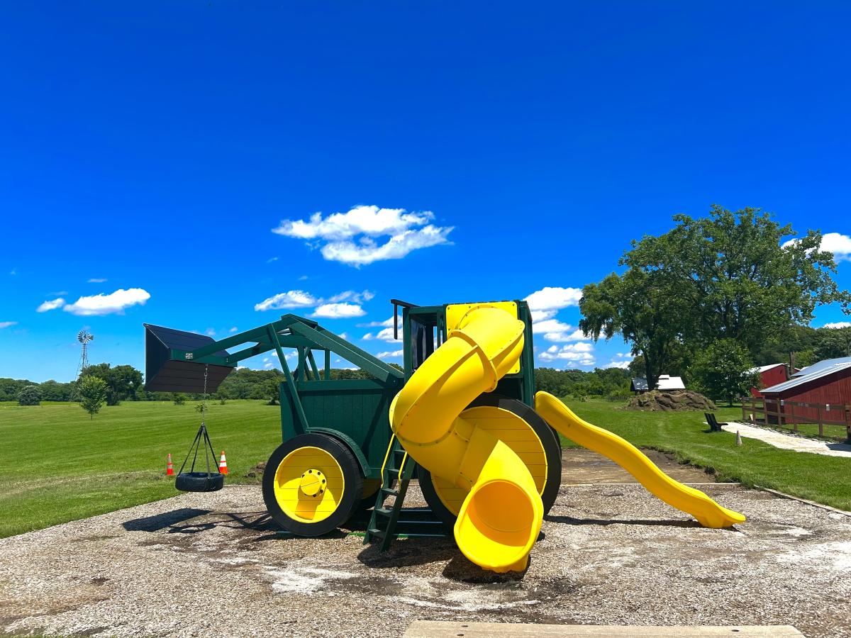 A green tractor-shaped playground with a yellow slide on a gravel surface under a bright blue sky.