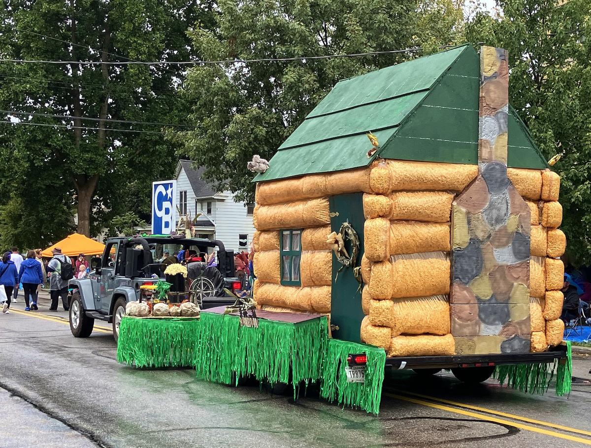 A float shaped like a log cabin is being pulled by a jeep.