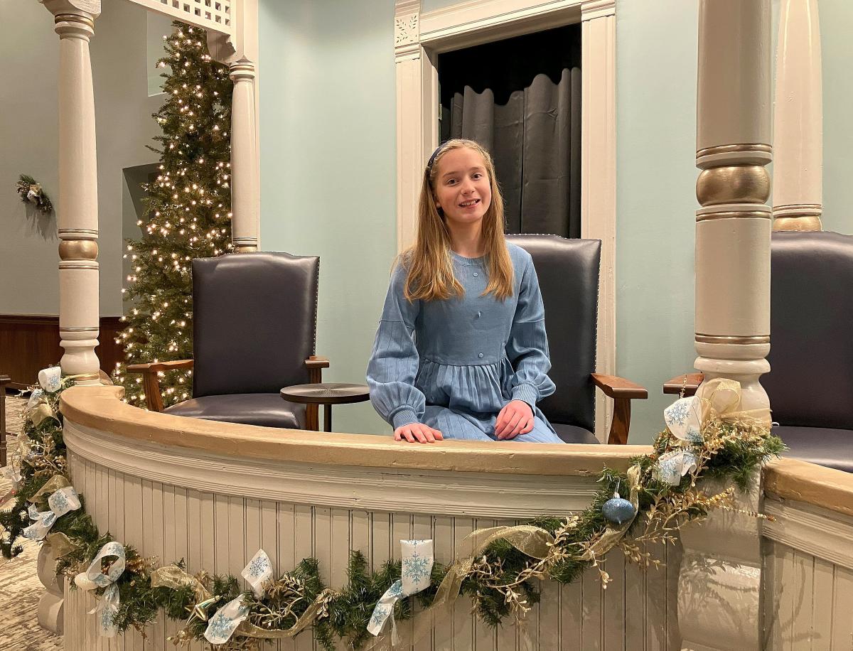 A young girl with long brown hair and a headband sits in an ornate theater box, smiling at the camera. She is wearing a long-sleeved blue dress and rests her hands on the wooden railing, which is decorated with festive garlands, gold ribbons, and blue ornaments. Behind her, two dark blue chairs are positioned near a black curtain, and a Christmas tree adorned with twinkling lights stands in the corner. The theater’s light blue walls and white columns add to the elegant setting.