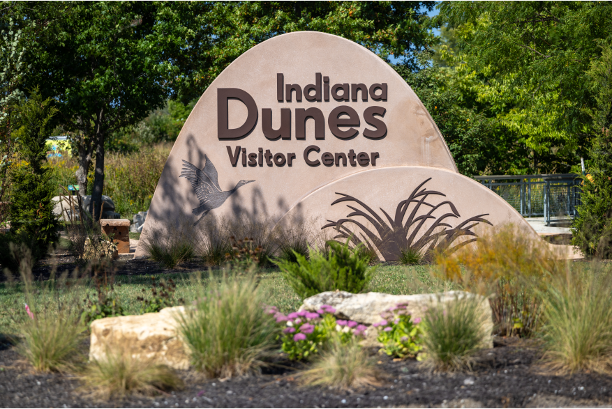 A dune-shaped sign says "Indiana Dunes Visitor Center" and has imagery of a sandhill crane and marram grass.