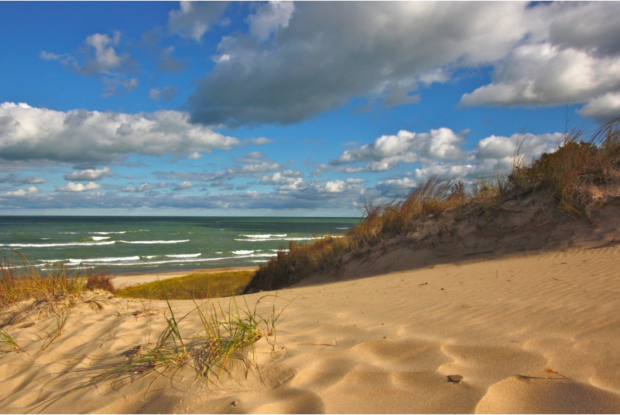 Sandy dunes with beach grass in the foreground and Lake Michigan visible under a partly cloudy sky at Indiana Dunes
