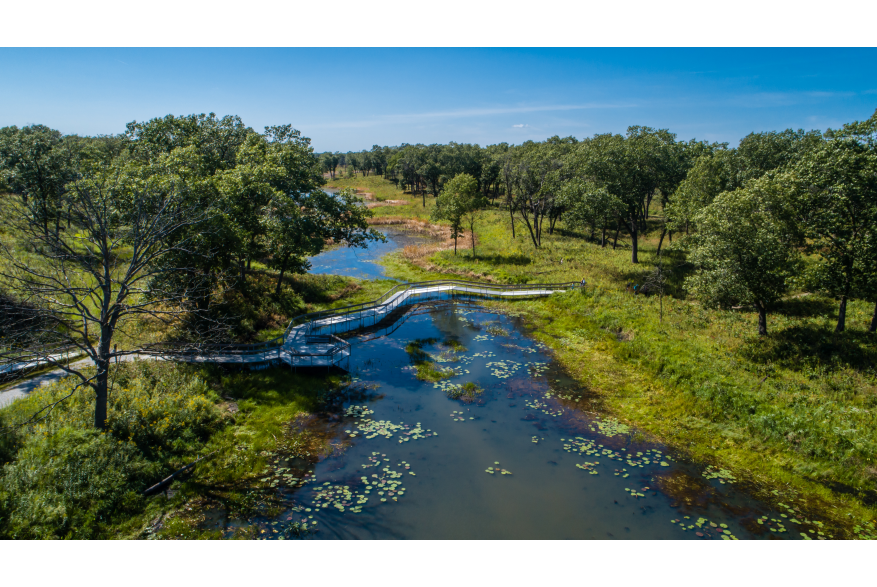 A boardwalk crosses over a waterway. Woods are seen on each side of the waterway.