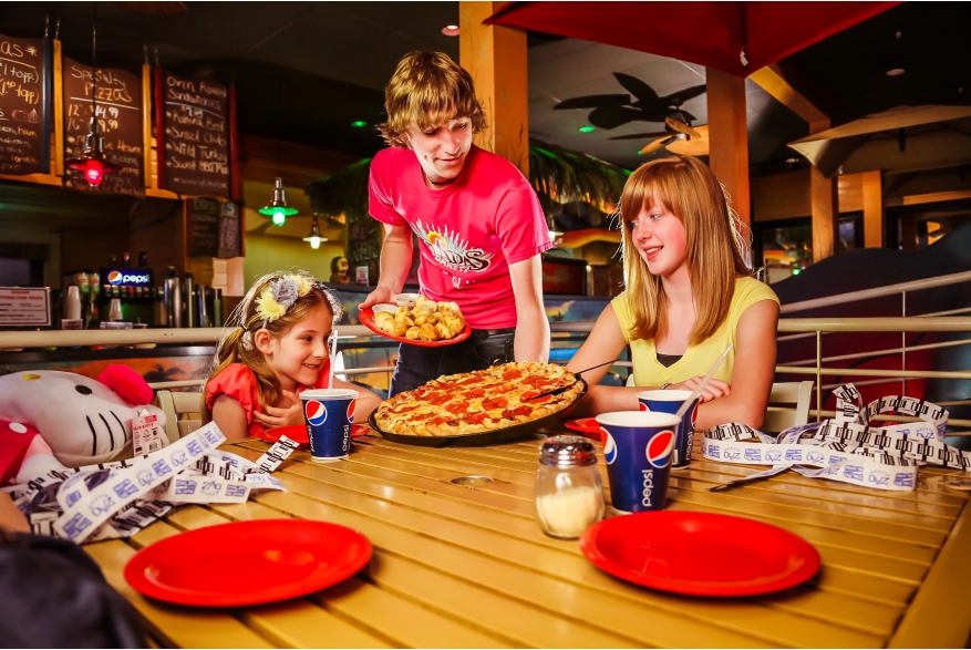 A waiter in a red shirt serves pizza to two young girls