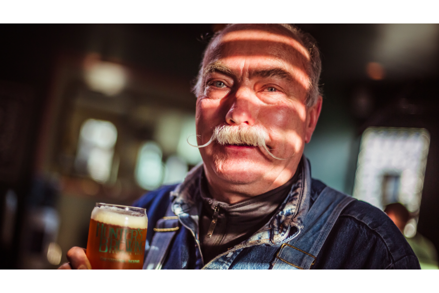 An older man with a curled mustache holds a glass of beer.