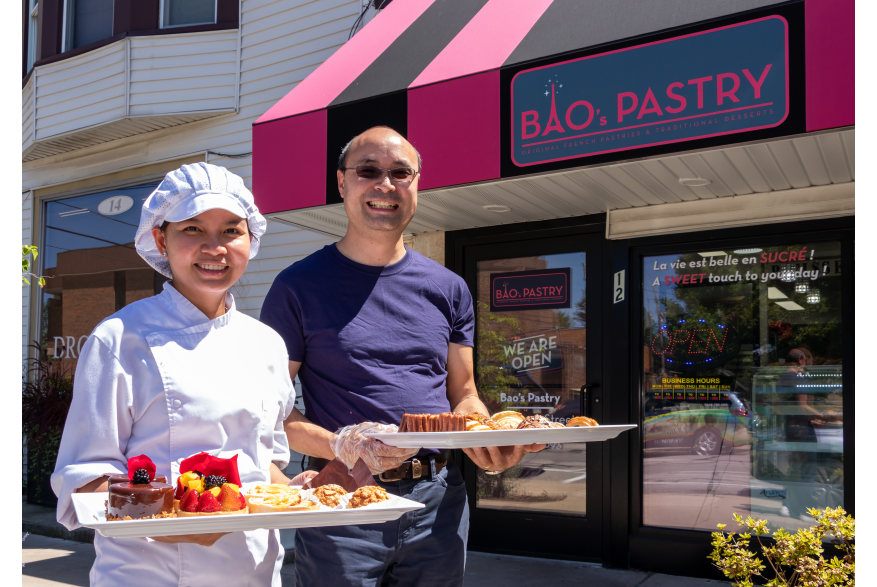 A chef and her husband stand in front of their pastry shop, holding trays of French pastries
