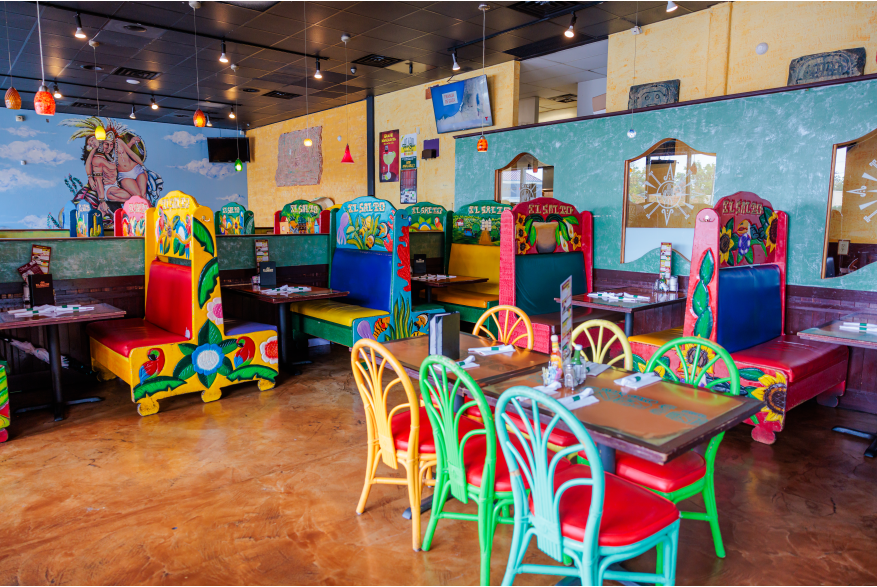 Colorful chairs, booths, and table are inside a restaurant. A mural and television are in the background.