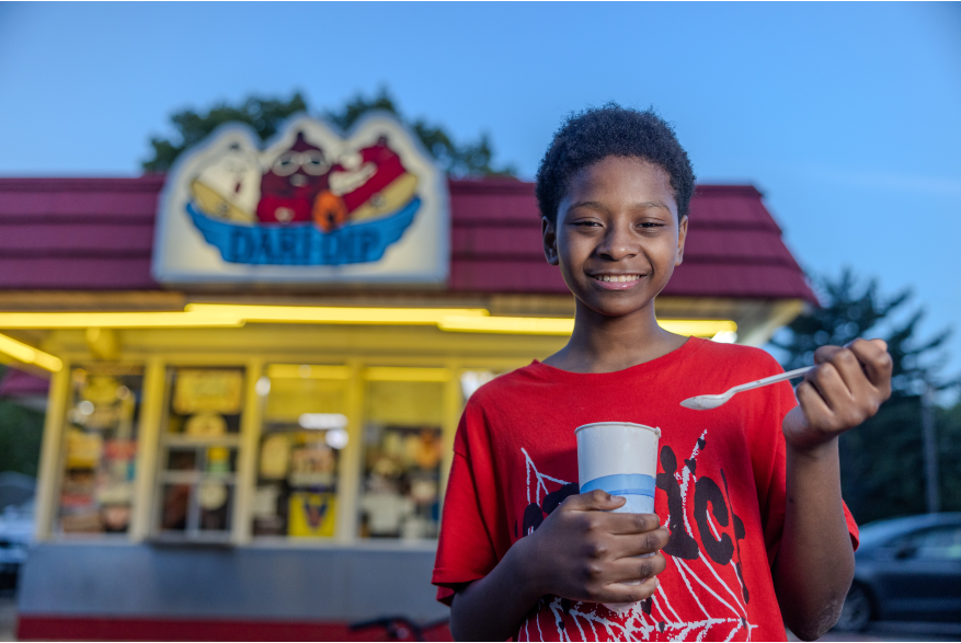 A young boy eats a shake in front of an ice cream stand.