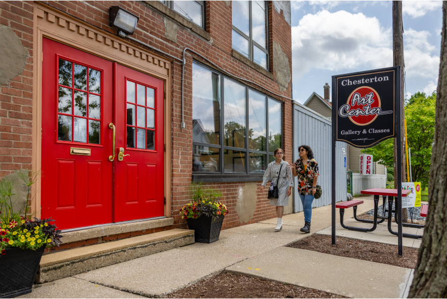 The exterior entrance of the Chesterton Art Center in Chesterton, Indiana, featuring striking red double doors set into a brick building with a gallery and classes sign. Two visitors walk along the sidewalk past potted flowers at the welcoming entryway.