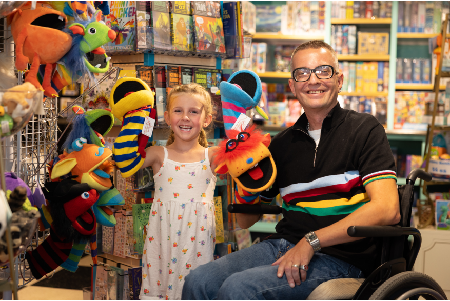 A young child stands next to a man sitting in a wheelchair. Both are smiling broadly and holding puppets.