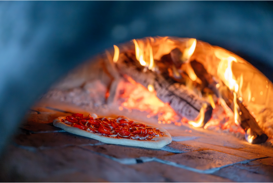 A pizza sits in a brick oven in front of logs that are aflame.