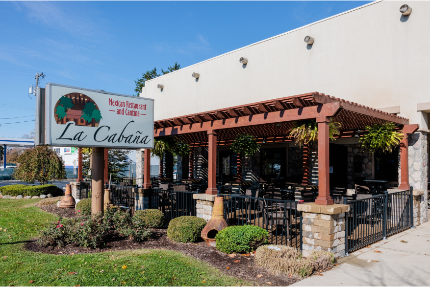 A stucco building with a fenced in patio sits near grass. The sky is blue. A sign out front reads La Cabaña and Mexican Restaurant and Cantina