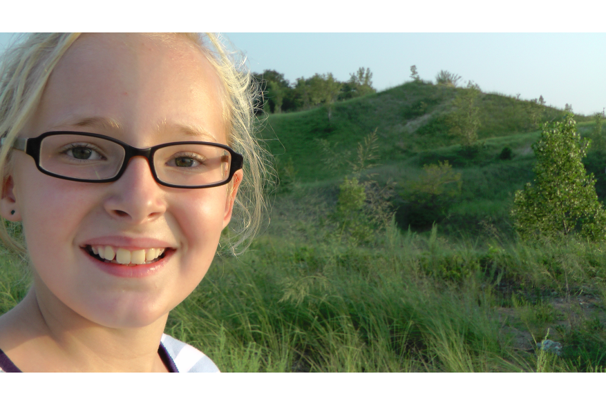 A blond girl with glasses is smiling. A vegetation-covered dune is in the background.