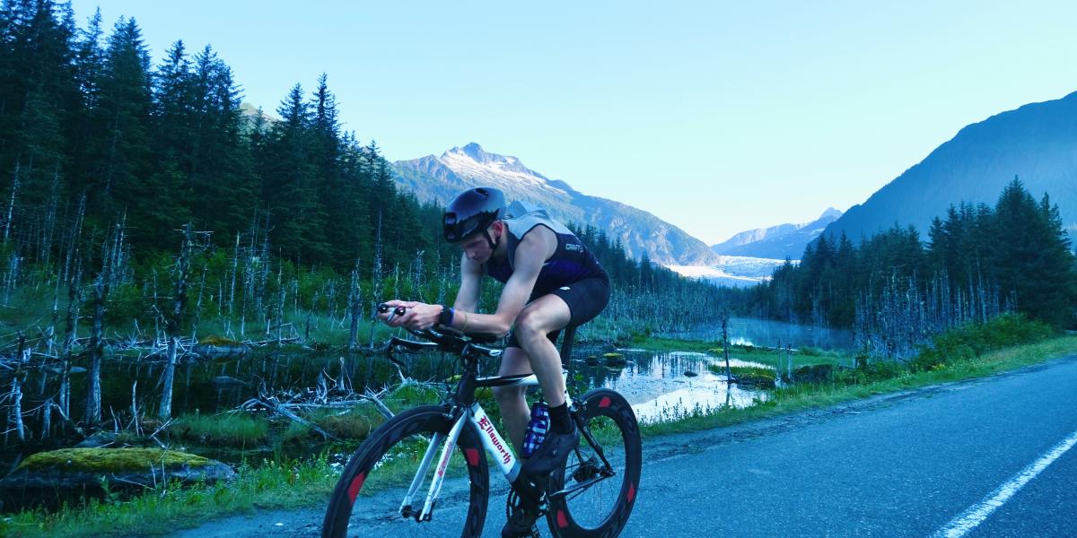 Photo of the bike racing portion of a triathalon racing down a road in front of the Mendenhall Glacier with mountains in the background and trees and a beaver pond in the foreground.