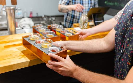 A person holds a wooden flight tray with multiple samples of colorful beverages at Electric City Ciderworks