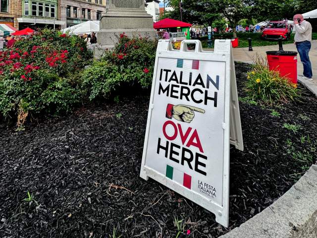 A sign for Italian Merch with a pointing finger, with the Electric City Sign and a statue in the background at La Festa Italiana