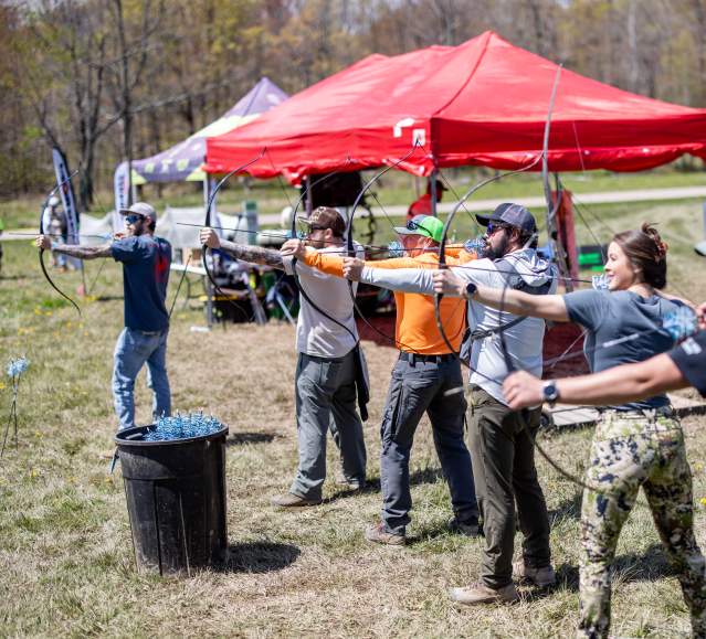 Archers line up and shoot during a festival at Camp Freedom, with event tents set up in the background.