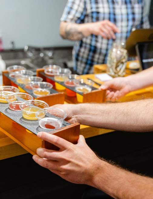 A person holds a wooden flight tray with multiple samples of colorful beverages at Electric City Ciderworks