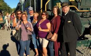 Women holding wine glasses and standing with a train conductor in front of a train