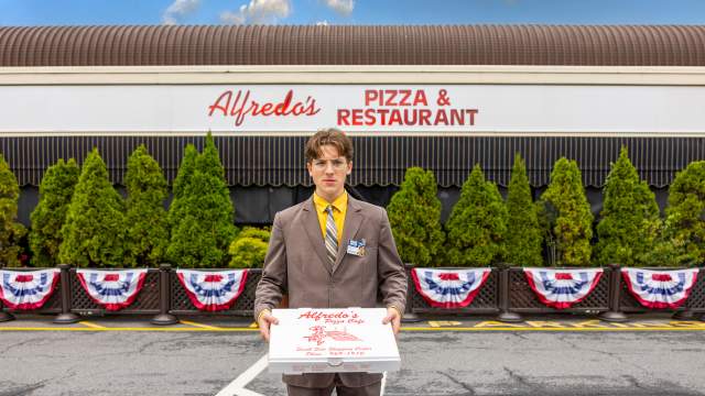 A man dressed up as Dwight Schrute is standing in front of Alfredo's Pizza Cafe while holding a pizza box