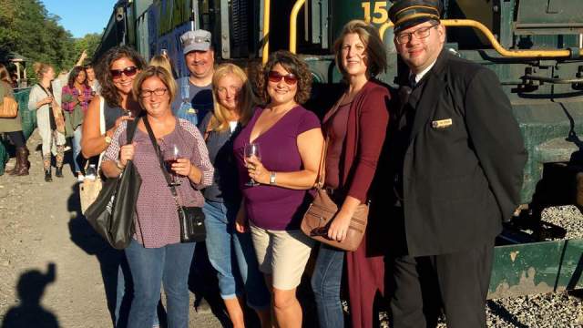 Women holding wine glasses and standing with a train conductor in front of a train