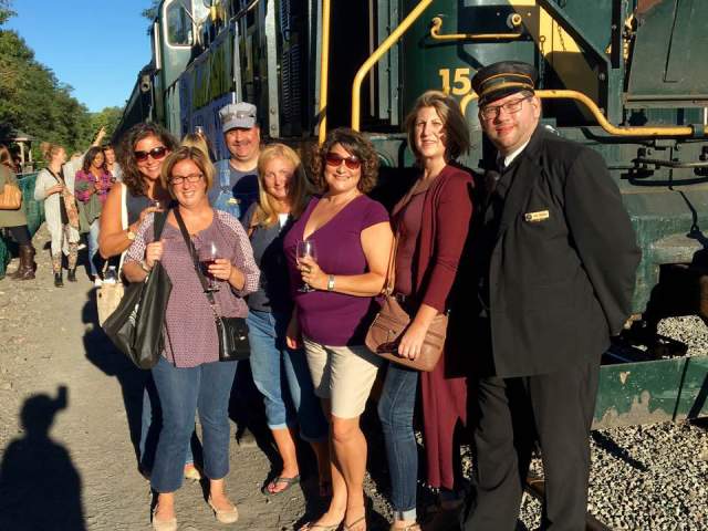 Women holding wine glasses and standing with a train conductor in front of a train