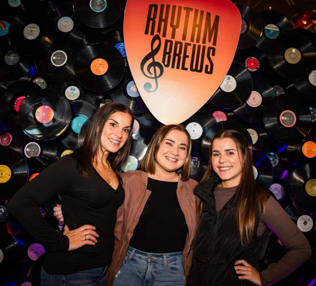 Three women posing for a photo in front of a Rhythm & Brews decorative wall featuring vinyl records at the Rhythm & Brews festival at Montage Mountain Resorts in Scranton, PA