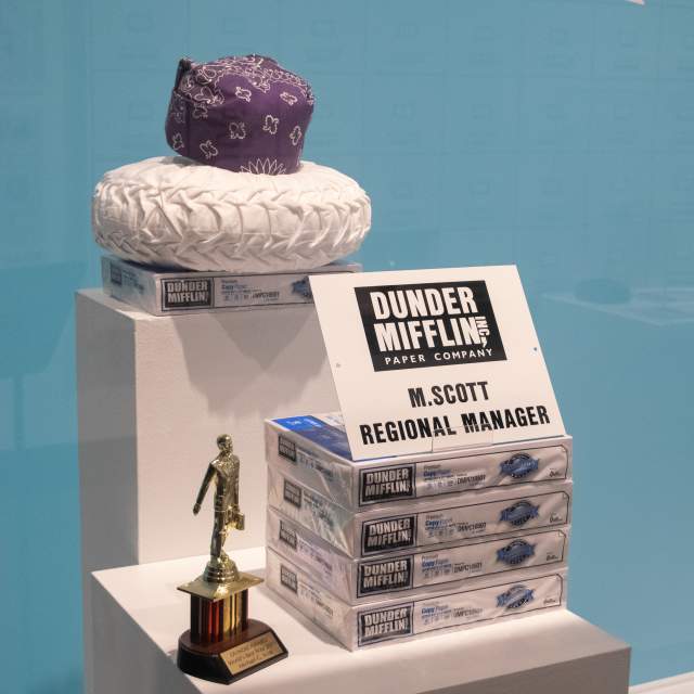 Props from The Office on display at The Office Exhibition at the Everhart Museum in Scranton, PA, including the iconic purple bandana, Dundie Award, and stacks of Dunder Mifflin paper.