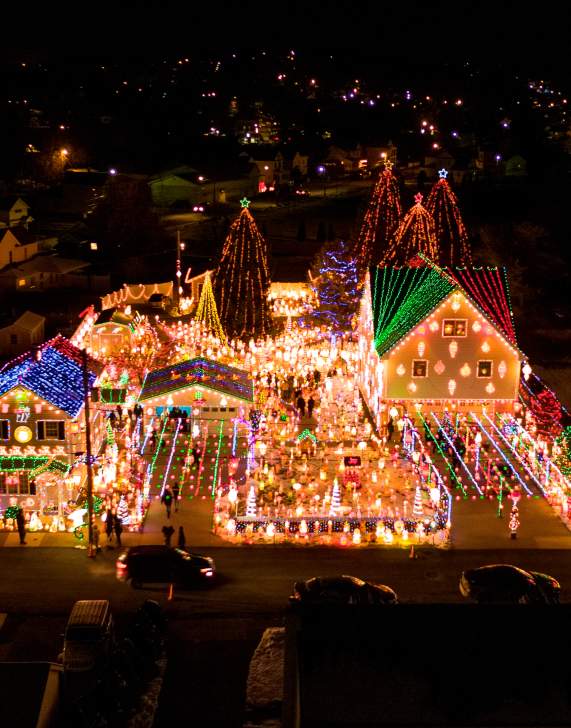 Aerial nighttime view of Peckville, PA neighborhood with colorful Christmas lights, decorated houses, and illuminated trees during the holiday season.