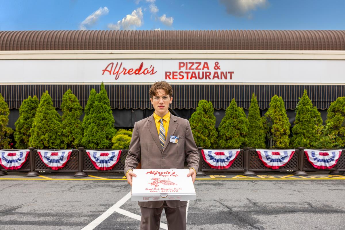 A man dressed up as Dwight Schrute is standing in front of Alfredo's Pizza Cafe while holding a pizza box
