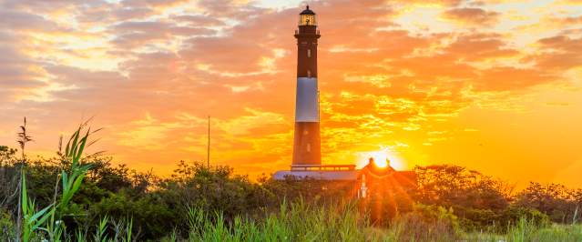 Tall lighthouse rising above coastal grasses on Long Island at sunset, with a glowing orange sky, scattered clouds, the sun near the horizon, and warm light illuminating the surrounding landscape.