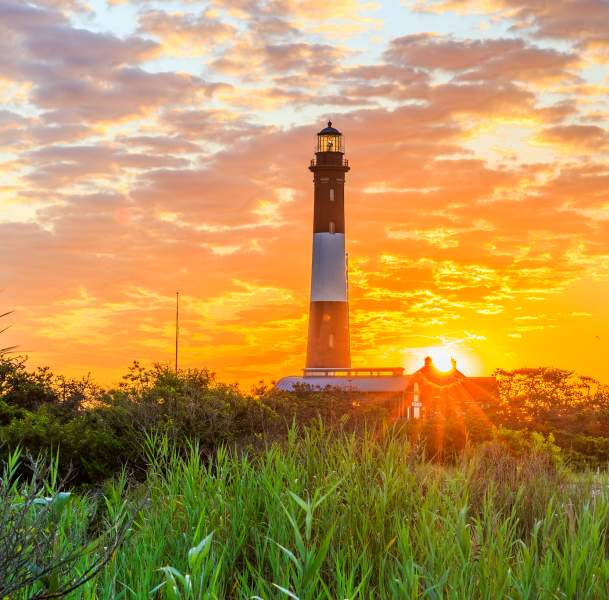 Tall lighthouse rising above coastal grasses on Long Island at sunset, with a glowing orange sky, scattered clouds, the sun near the horizon, and warm light illuminating the surrounding landscape.